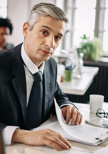Portrait mid adult businessman sitting desk going through business reports while looking camera office