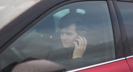 View young male wearing black jacket sitting red car while talking phone