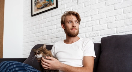 Young man smiling stroking cat sitting sofa home