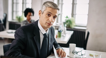 Portrait mid adult businessman sitting desk going through business reports while looking camera office