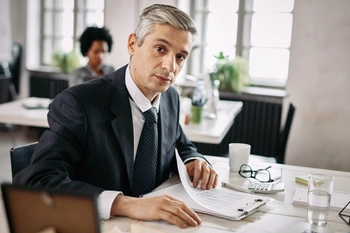 Portrait mid adult businessman sitting desk going through business reports while looking camera office