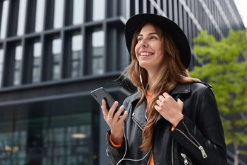 Photo charming dreamy woman holds modern cellular listens music walk outdoor metropolis