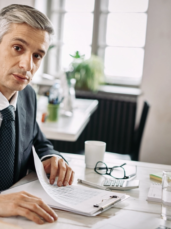 Portrait mid adult businessman sitting desk going through business reports while looking camera office