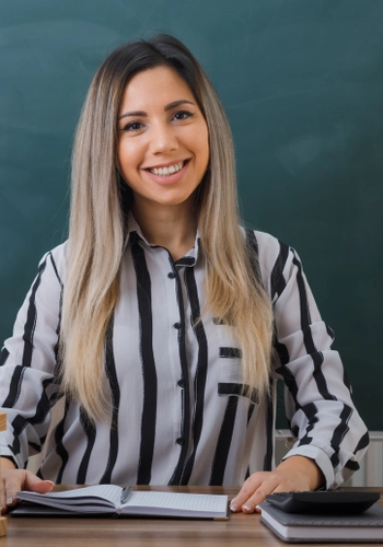 Young woman teacher sitting school desk front blackboard classroom checking homework students looking camera happy pleased smiling