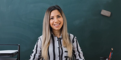 Young woman teacher sitting school desk front blackboard classroom checking homework students looking camera happy pleased smiling
